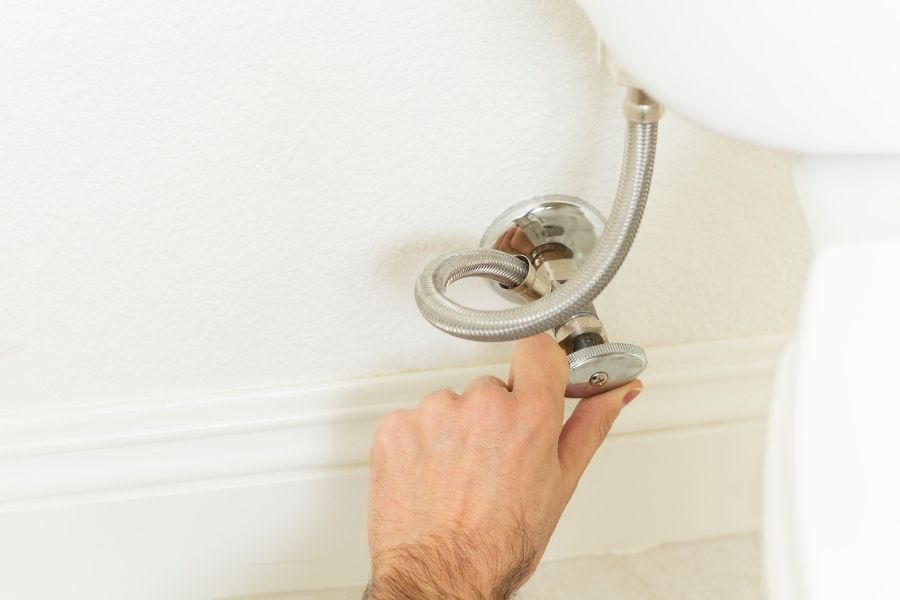 A hand reaching behind a toilet to turn the water shutoff valve clockwise to stop an overflow
